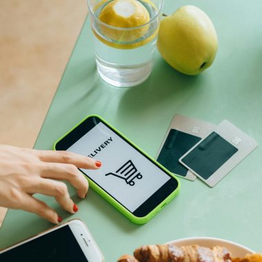 Hand using mobile app for food delivery. Croissant, apple, and credit cards on table.